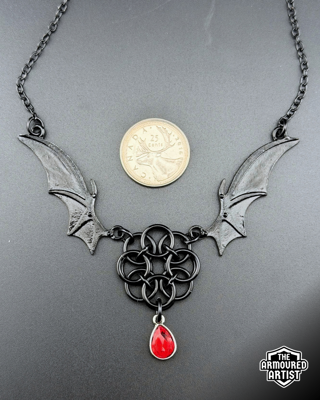 A gothic-style black metal necklace featuring bat wings, a round chainmail knotwork center, and a dangling red teardrop gem, displayed on a black background with a Canadian quarter for scale.