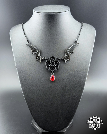 A gothic-style black-toned necklace displayed on a black bust. The necklace features bat-wing motifs on either side, connected by a central interwoven knot design, with a dangling red teardrop gemstone.
