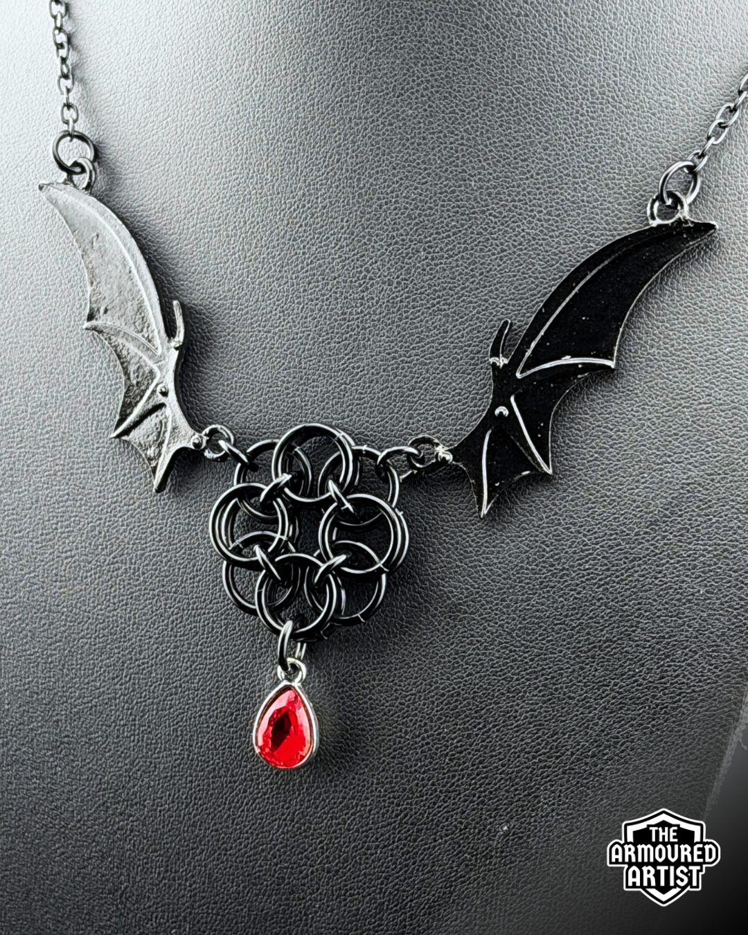 A gothic-style black-toned necklace displayed on a dark gray bust. The necklace features bat-wing motifs on either side, connected by a central interwoven knot design, with a dangling red teardrop gemstone.