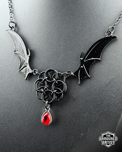 A gothic-style black-toned necklace displayed on a dark gray bust. The necklace features bat-wing motifs on either side, connected by a central interwoven knot design, with a dangling red teardrop gemstone.