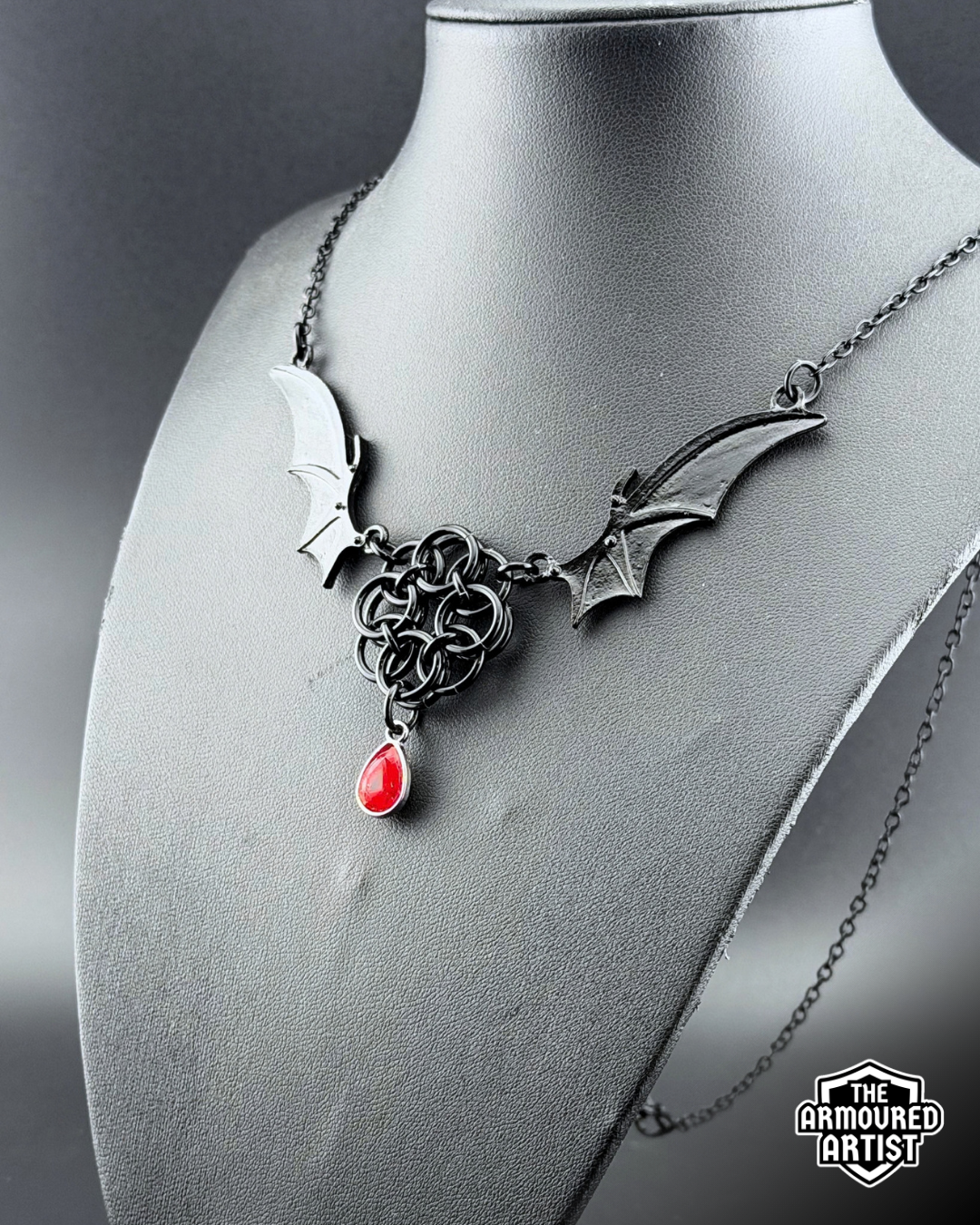 A gothic-style black-toned necklace displayed on a dark gray bust. The necklace features bat-wing motifs on either side, connected by a central interwoven knot design, with a dangling red teardrop gemstone.