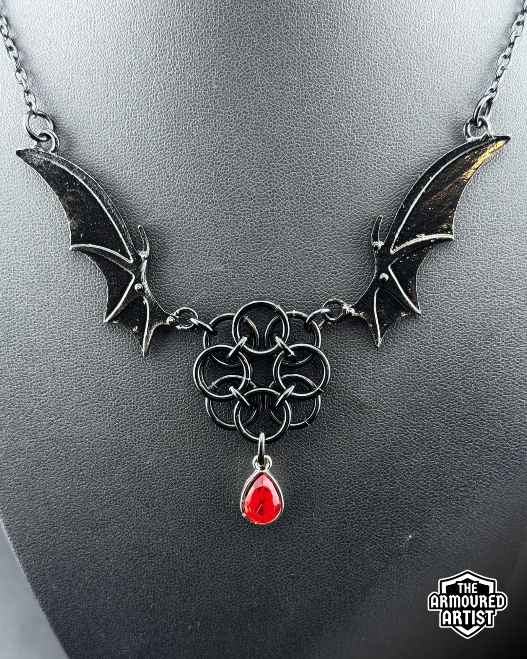 A gothic-style black-toned necklace displayed on a dark gray bust. The necklace features bat-wing motifs on either side, connected by a central interwoven knot design, with a dangling red teardrop gemstone.